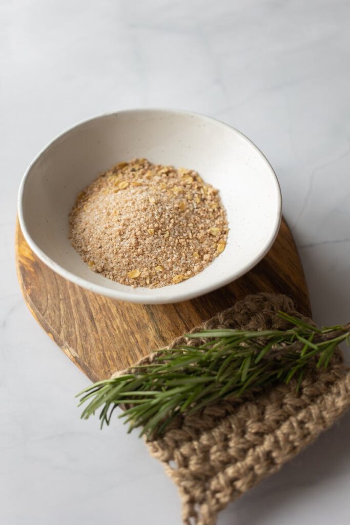 white bowl filled with powder of oat flakes near fresh verdant branch of rosemary on timber board